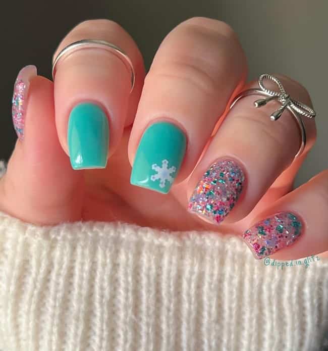 A closeup of a woman's mid-length nails with teal and blue and pink glitter nail polish that has white snowflakes on teal nails 