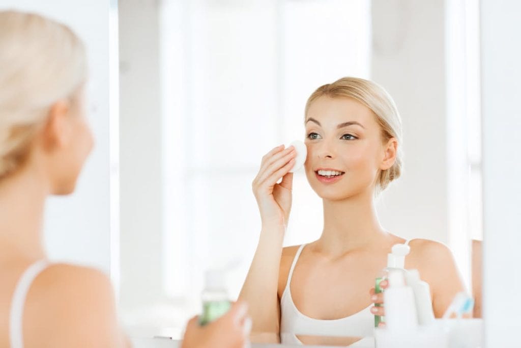 a blonde woman applying toner on her face using a cotton pad while looking at the bathroom mirror