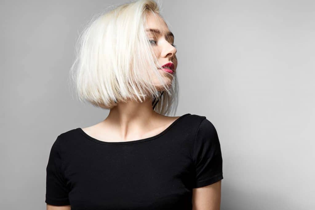 A stunning blonde woman with platinum hair dye, wearing a black top and posing against a gray background.