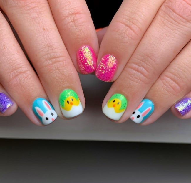 A woman's nails are decorated with two glittery bright pink and purple nails and two green and blue accent nails with bunny and chicks