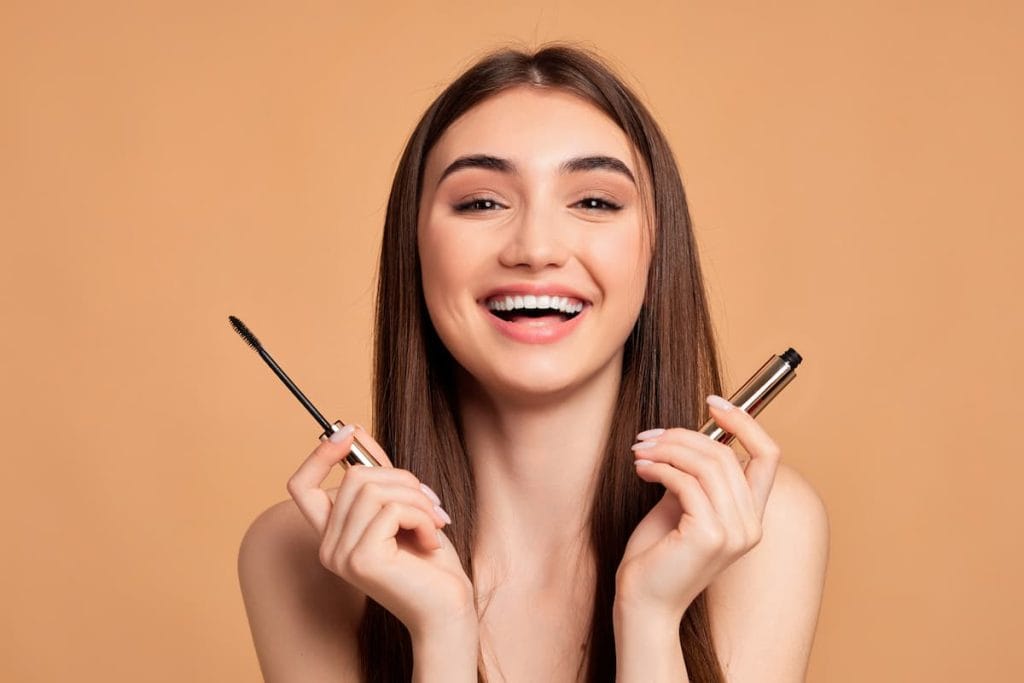 A laughing young woman holding a mascara on a beige background.