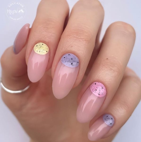 A woman's nails mani features half-moon designs in chic yellow, purple, and pink colors with speckled egg patterns on a nude base