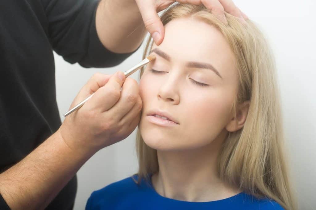 a make up artist applying concealer around the eyebrow of a blonde young woman using a brush