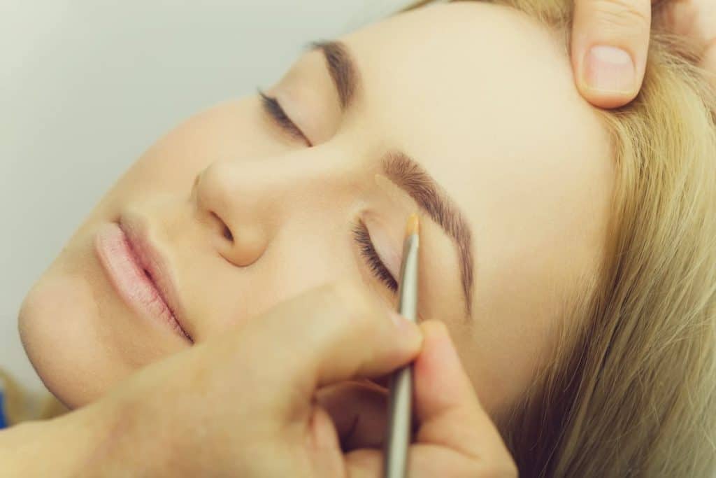 a close-up image of a woman with her eyes closed while a makeup artist is applying eyebrow concealer