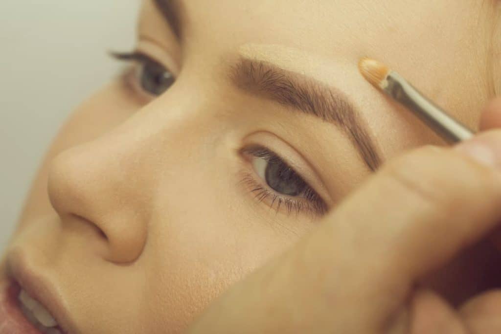 a close-up image of a make up artist putting concealer on her client upper brows