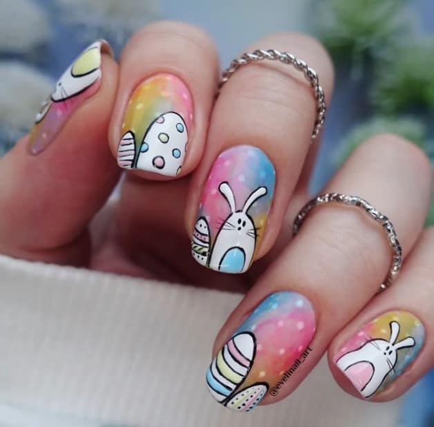 A woman's nails with watery mix of bright colors like blue, pink, green, and yellow, serving as the perfect backdrop for cute bunnies and Easter eggs with stripes and polka dots