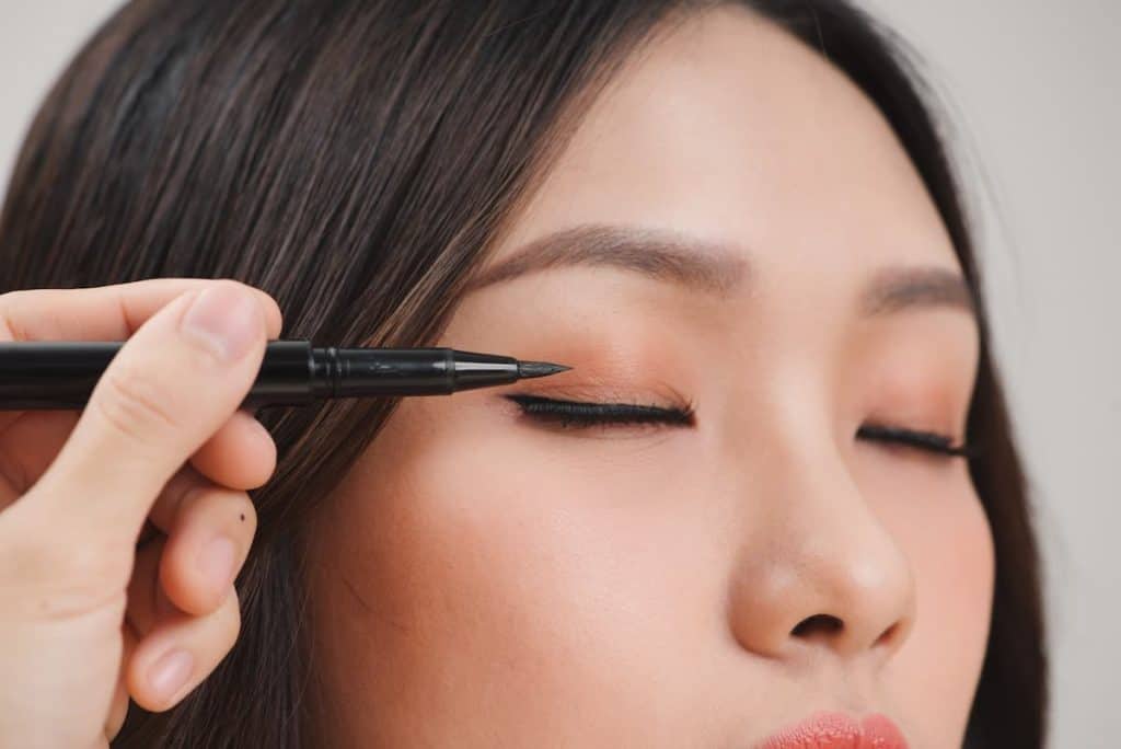 A young asian woman with her eyes closed while a woman is using a black eyeliner on her eyes.