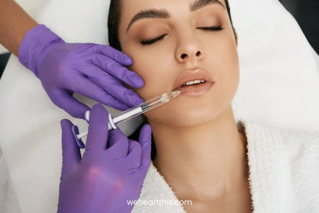 a woman laying down while wearing white robe and a professional with purple gloves is injecting a lip filler on her lower lip
