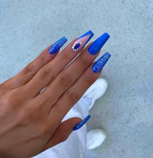 A woman's hand with glossy royal blue done up in glitter, V tips, and studded with diamonds on an accent nail