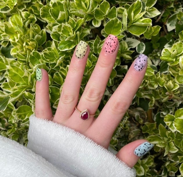 A woman's hand which features pastel yellow, pink, green, and purple dip nails that serve as the base for black speckles