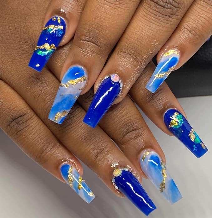 A woman's nails with glossy blue nails with marble accent nails over a clear base, topped with stones at the cuticle and gold foil