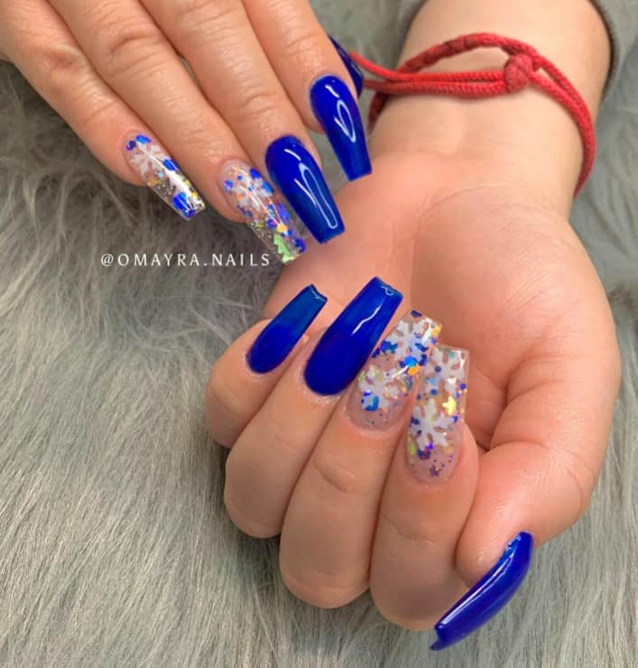 A woman's hands with gloss and translucent blue accent nails sprinkled with encapsulated Christmas ornaments like snowflakes, stars, and pine trees