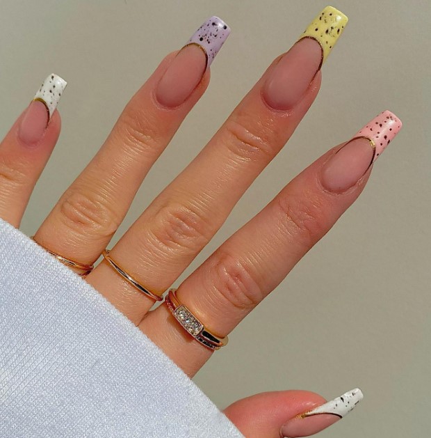A woman's hand features white, yellow, pink, and purple French tips with speckled patterns and gold outlines on nude coffin nails