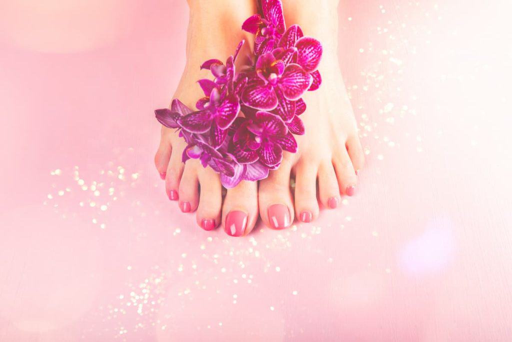 A woman's feet adorned with purple orchids and decorated using acrylics for toenails on a pink background.