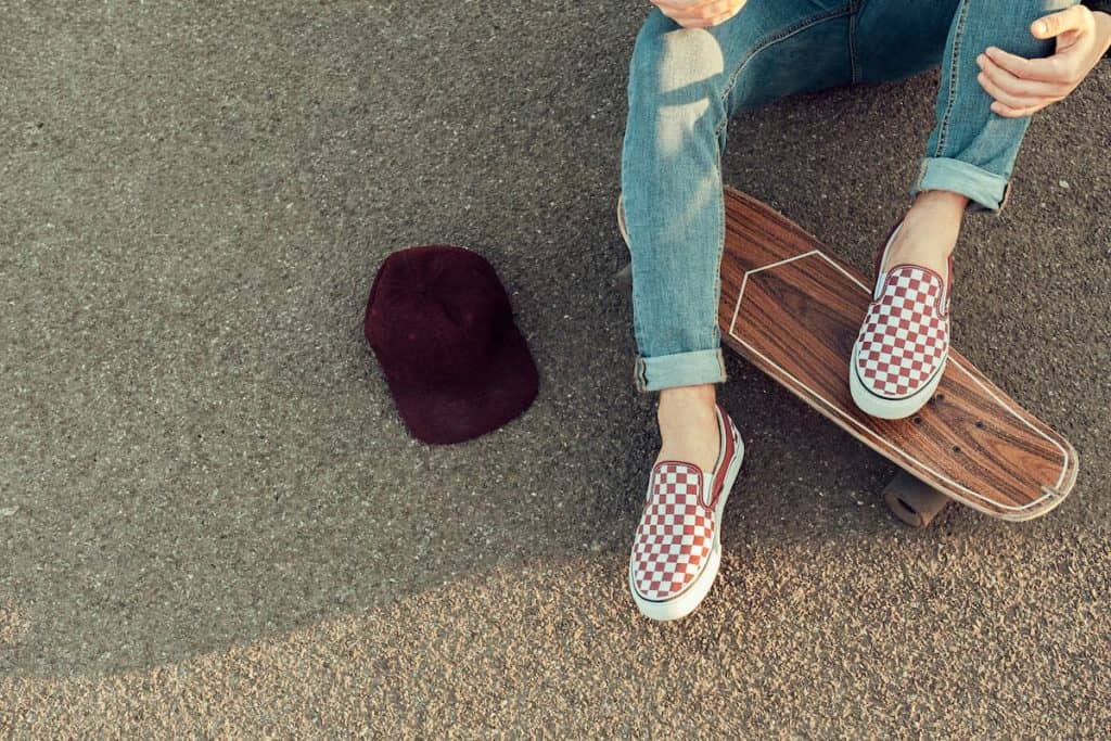 A person sitting on a skateboard wearing checked vans sneakers with his hat beside him
