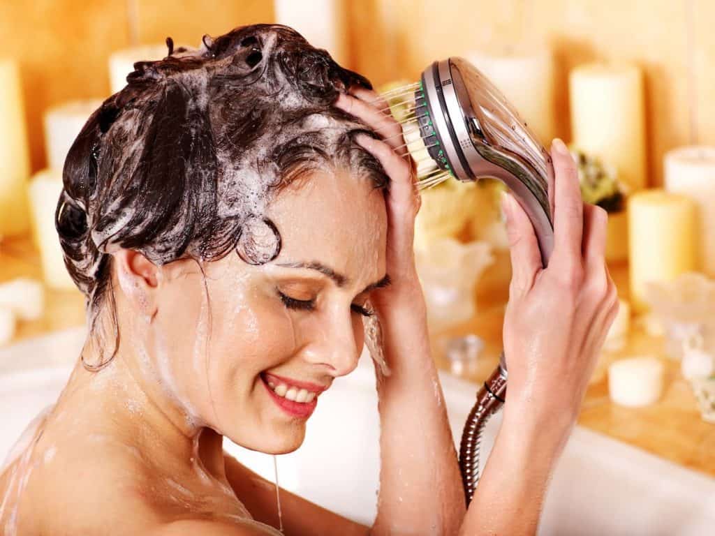 A woman is washing her hair with a shower head while sitting in a bathtub.