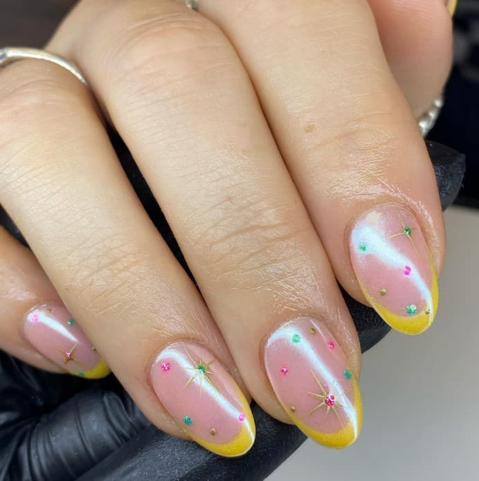 A woman's hand with nails feature gold and glittery stars with 3D rhinestone centers on a light pink base with stones in various colors and lemon-yellow half-moon tips