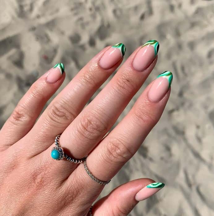 A woman's hand with nude nails and French tips with swirls of shades of light to dark green on the beach