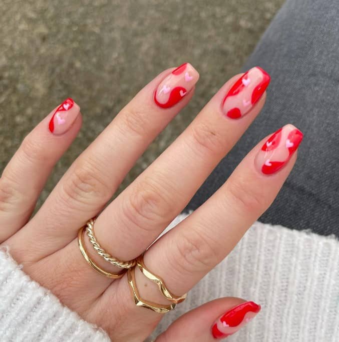 A woman's pink and red nail design features red swirls painted randomly near the tips, sides, and cuticles against a nude base and decorated with tiny pale pink hearts