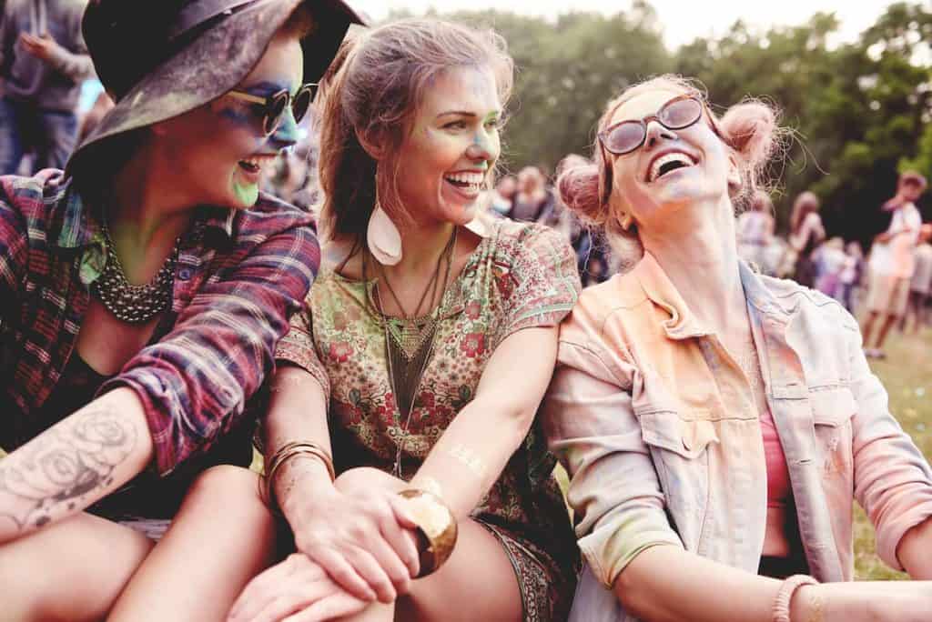 Three women sitting on the grass at a music festival happily flaunting their concert outfits and accessories