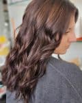 The back of a woman's hair wearing a gray shirt with long wavy hair and tansel.