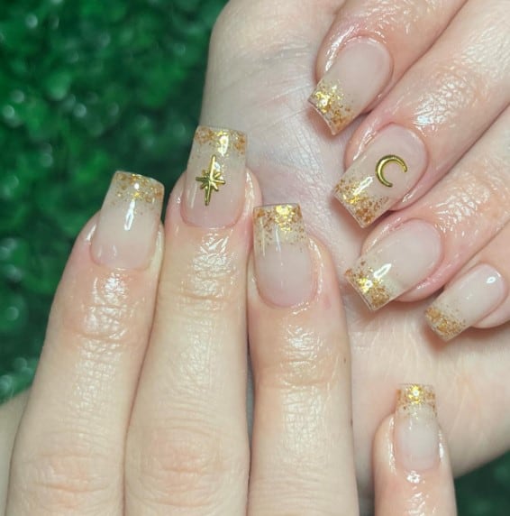 A woman's nails are decorated with specks of gold foil on your nail tips and using gold nail polish to create a star and a crescent moon design on both middle fingers