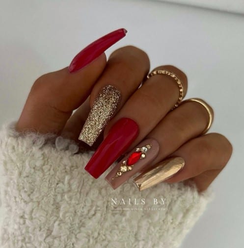 A woman with gold chrome nails with gold glitter, shiny red nails, and a nude-colored one with stunning red and gold rhinestones