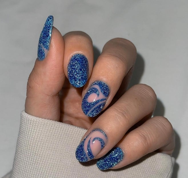 A woman's mid-length oval nails are painted in soft peach and adorned with dainty swirls of fine blue glitter