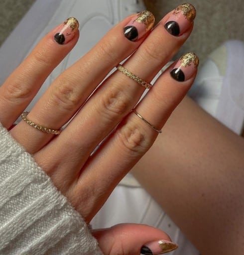 A woman's hand with black half-moons on your nail beds with glittery gold French tips over nude nails