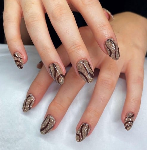 A woman's nails are dipped in rich gold glitter and adorned with playful brown swirls