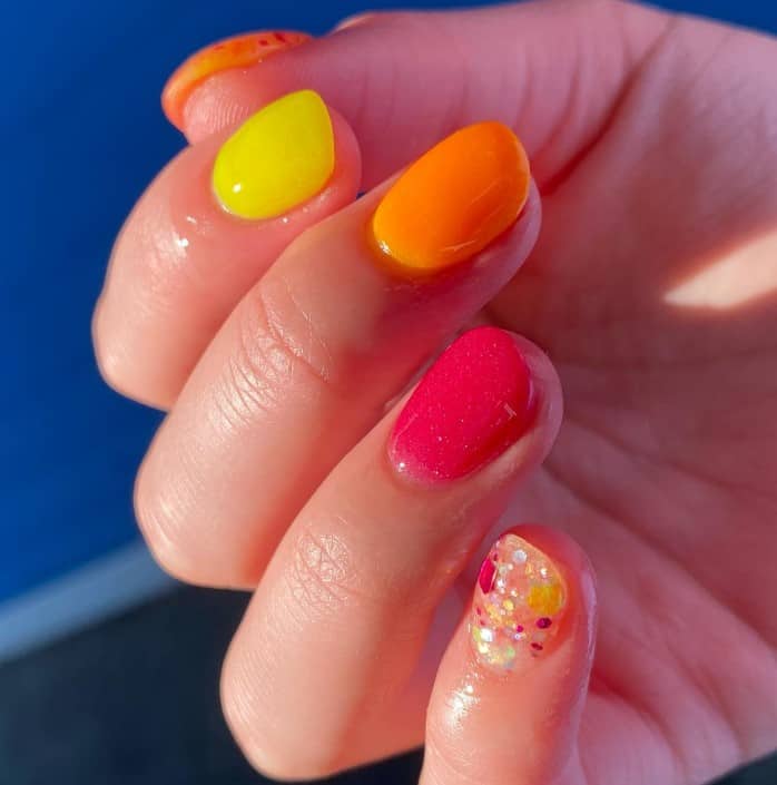 A person holding up a brightly colored manicure with encapsulated accent nails with multicolor splashes and chunky glitter.