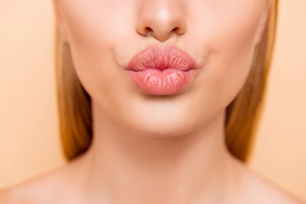 A woman is making a kiss with her lips painted with peach colored lipstick.