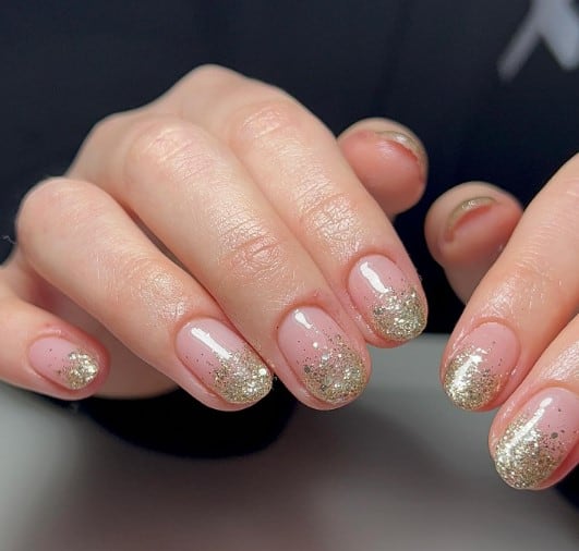 A woman's short peach nails shimmer with tips dipped in luxurious gold fine glitter