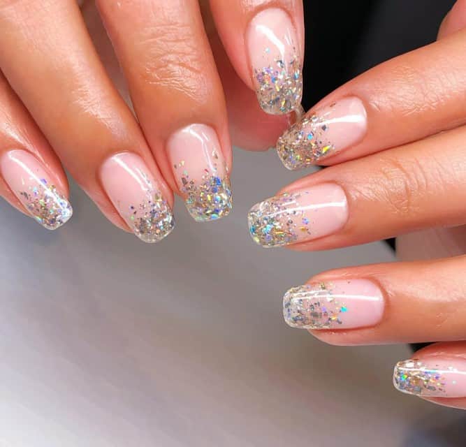 A woman's long squoval nails glow in soft peach and take a playful twist as their tips become confetti-like bursts of silver