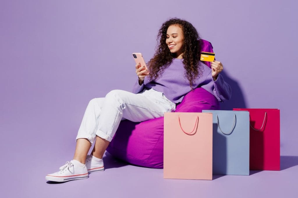 Full body young woman wear pullover sit in chair near package bags after shopping use mobile cell phone credit card isolated on plain pastel light purple background.