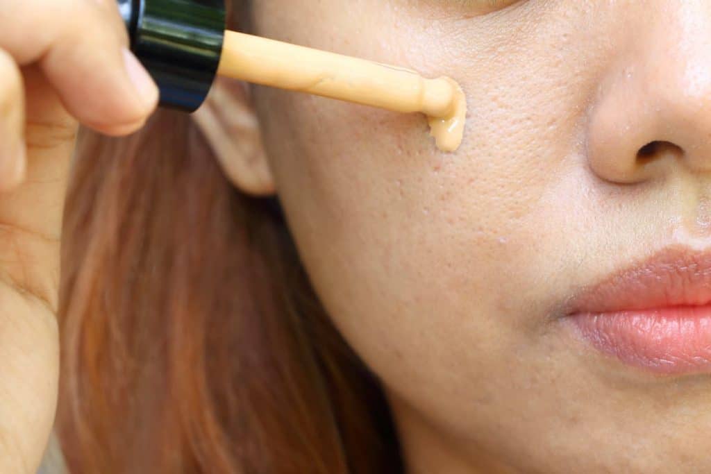 Closeup the Asian woman use pipette drop cosmetic liquid foundation on her cheek skin for hide acne and wide pore on face skin before makeup.