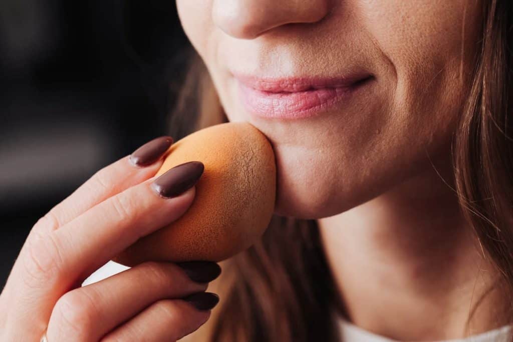 a close up image of a girl applying foundation on her face using an orange sponge