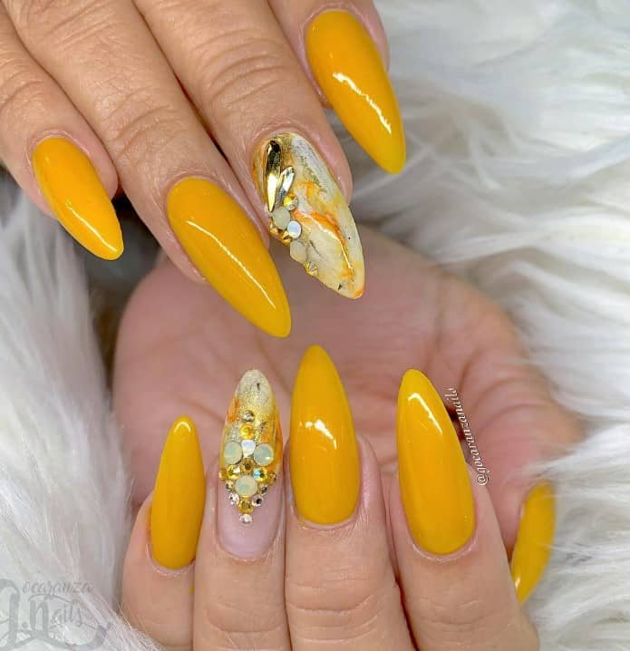 A woman's hands with mustard yellow nail design with nude accent nails embellished with yellow, gold, and gray marble effects and completed with gold and silver 3D stones for a luxe finish