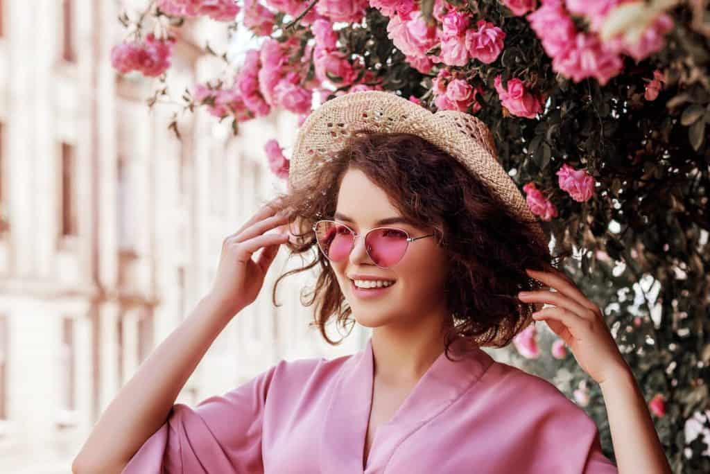 Outdoor close up portrait of young beautiful happy smiling curly girl wearing stylish pink sunglasses, straw hat, dress.