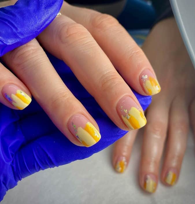 A woman's nails features nude base with paint strokes in varying shades of yellow coming down half the nail and finished with accents of holographic crackle foil that shine as gold, light blue, and silver depending on the light