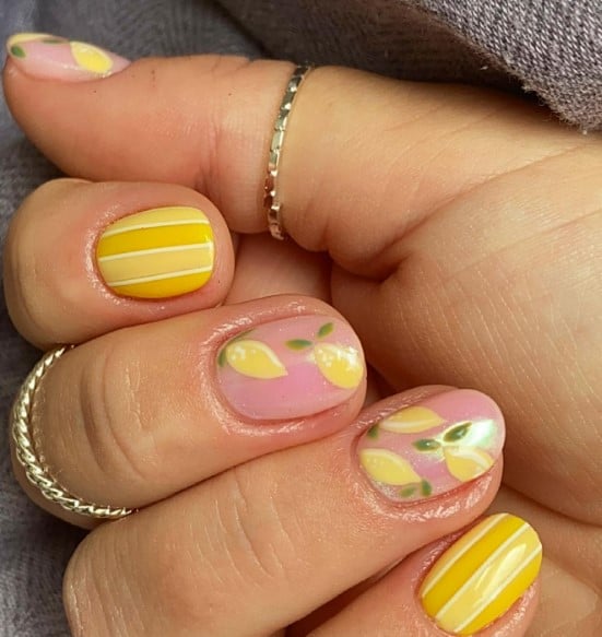 A woman's hand features a two-toned yellow base with light yellow vertical stripes and nude accent nails decorated with lemon nail art