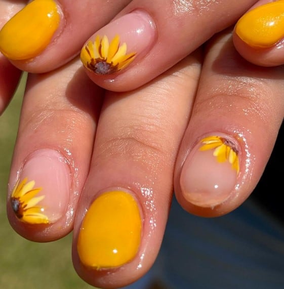 A person's nails with yellow set with nude accent nails that feature sunny sunflowers peeking from the tips and the base of different nails