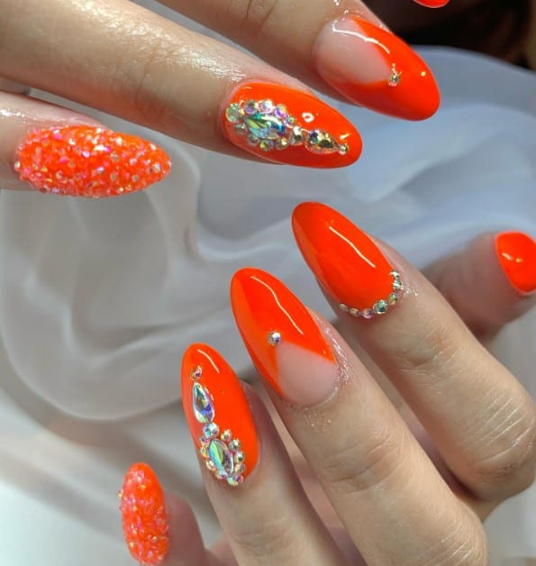 A woman's bright orange nails with glitter and jewels in different sizes