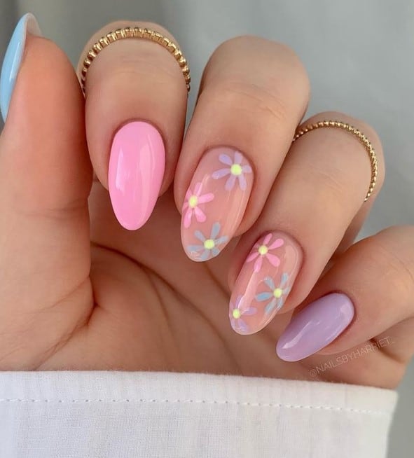 A hand with pink, purple and blue pastel nails and accent nails with delicate floral designs in the same hues, featuring a vibrant yellow center on a neutral base