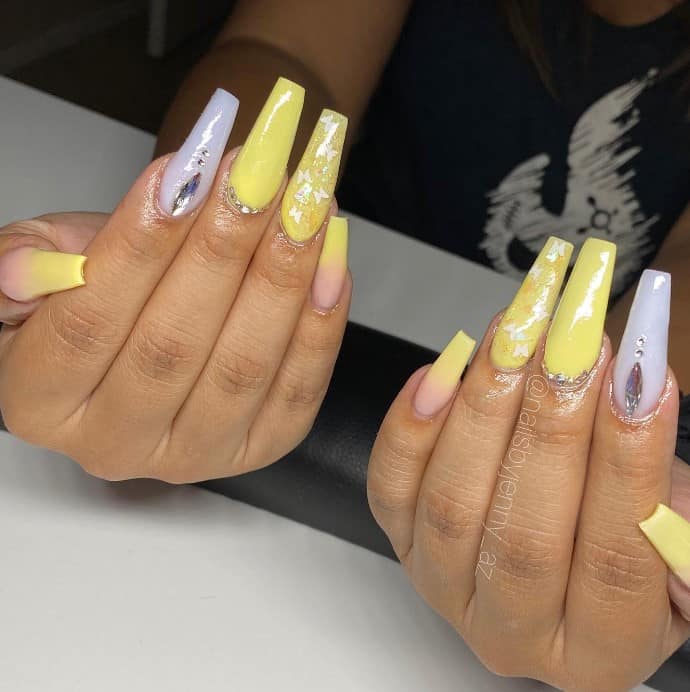 A woman's hands features light yellow set with silver rhinestones around the cuticles and silver butterfly nail art for multidimensional yellow coffin nail designs