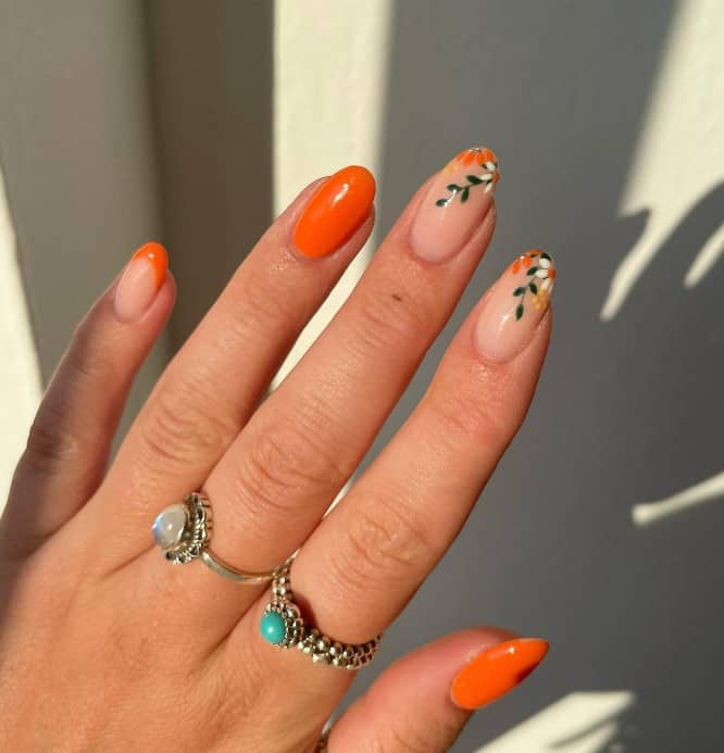 A woman's hand mani showcases bold orange nails, one nail with an orange French tip, and two nails tipped with orange floral designs in summery colors like white, green, and yellow
