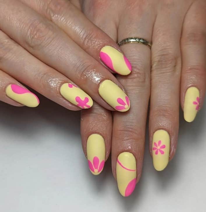 A woman's nails features a matte pastel yellow base with various pink shapes as nail decor, including flowers, stars, and swirls.