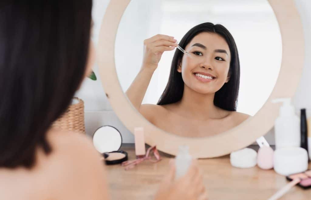 Happy beautiful asian girl applying eye serum in front of mirror at home