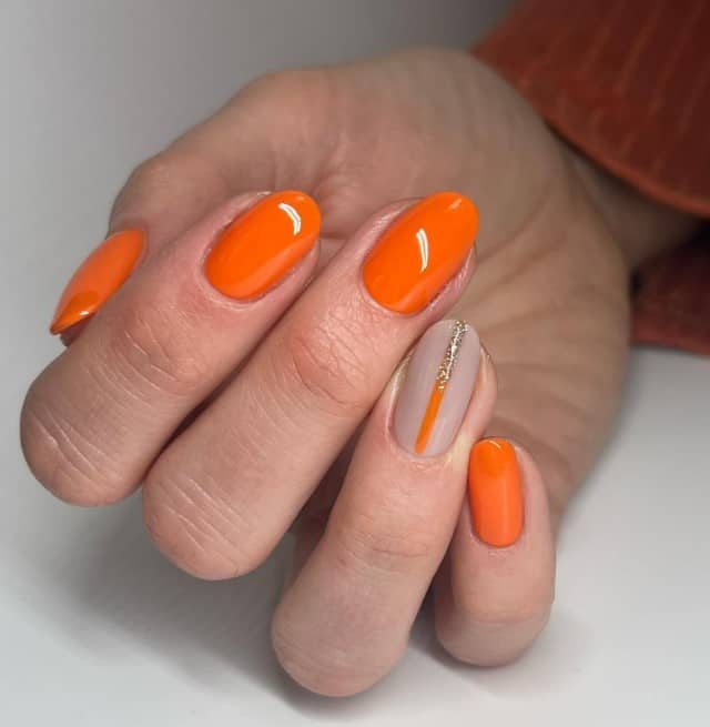 A woman's hand mani features bold and glossy orange nails with a nude accent nail decorated with a thin half-orange, half-gold glitter line down the middle