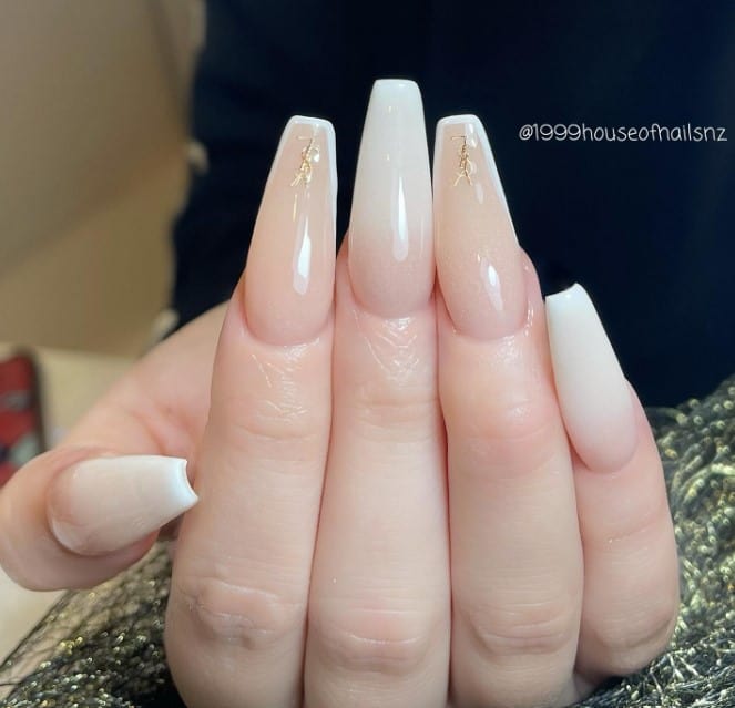 A woman's hands showcasing nude and off-white ombré nails dazzle with a glittery finish, while golden YSL logos add a luxurious touch to the subtle white French tips.